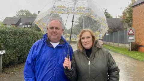 A man in a blue coat and a woman in a green coat standing under an umbrella. The road behind them is flooded. 