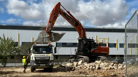 Mousumi Bakshi/BBC A red digger is picking up a pile of rubble and putting it into a white lorry, which a man in a hard hat and a hi-vis jacket is standing next to. In the background is a large white building.