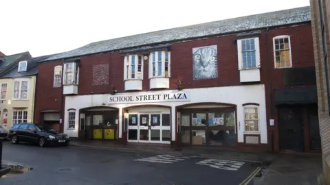 Exterior or School Street Plaza - a two-storey brick building in a row of terraced houses. The frontage takes up the space of about three or four terraced houses. There are double doors in the centre and two units either side with their own entrances on to the street. The whole building is abandoned with posters and newspaper stuck over the windows.