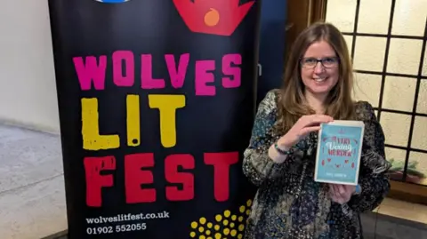 Lucy Andrew A woman holding up a blue paperback with A Very Vexing Murder written on the front. She has long brown hair and is smiling. She is wearing a blue and green patterned tunic. She is standing next to a black banner which says Wolves Lit Fest.