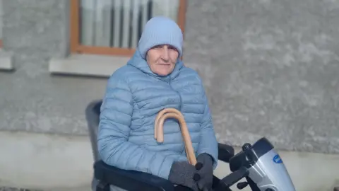  Ellen Shells sits in a mobility scooter outside a grey rendered building.  She is wearing a light blue beanie hat, a light blue puffed jacket and grey woolen gloves.  She is holding two wooden walking sticks.