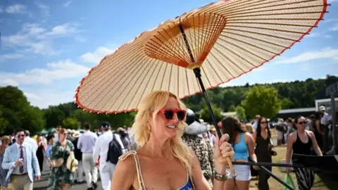 A woman holding a parasol and wearing red sun glasses, with crowds at the Henley Regatta behind her