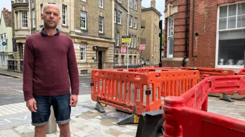 A man standing on the street beside some bright orange pavement works, with his business in the background.