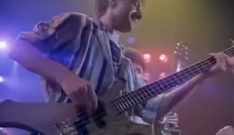 PA Pixely screengrab from an old music video, which shows a man in a shirt playing a bass guitar with bright lights behind him