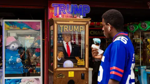 Emma Warley A man stands looking into an arcade in Scarborough. He is holding an ice cream and wearing a baseball top. He is looking at a 'truth teller' box which has a figurine of Donald Trump inside. A faint reflection of Warley, taking the shot, can be seen in a window.