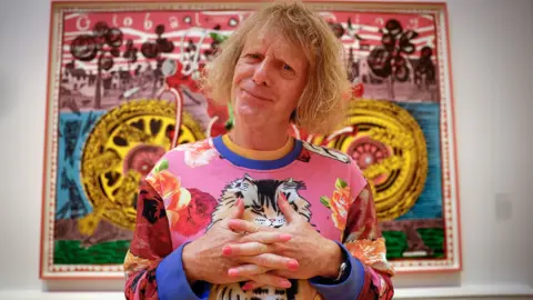 Grayson Perry folds his hands while posing in front of his painting