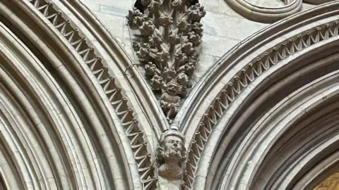 Eleanor Maslin/BBC The stonework inside a cathedral. Two arches move away from each other and in the middle is a head carved into the stonework. Above it is an imp - a little creature with its mouth open - and above that is a carved display resembling flowers.