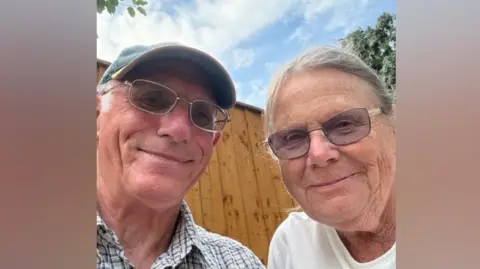 A selfie-style image of an elderly man and an elderly woman. They are wearing glasses and smiling while looking into the camera. The man is wearing a baseball cap. They are both wearing glasses. 
