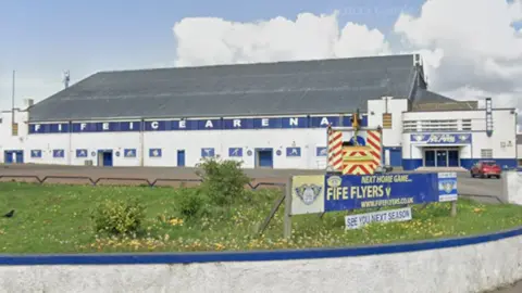 Google Fife Ice Arena is a white building with a grey roof. It has FIFE ICE ARENA on white letters along blue panels on the wall just below where the roof begins. It has several blue doors and a car park in front of it, with grass in the foreground. A sign by the entrance to the car park reads "Next home game... Fife Flyers v" with a blank space.