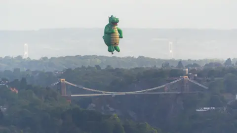 Sam Binding A giant green hot air balloon in the shape of a dragon flies high above Clifton Suspension Bridge. It has a yellow belly and a smiling mouth as well as two small horns. The Prince of Wales bridge is just about visible in the far distance.