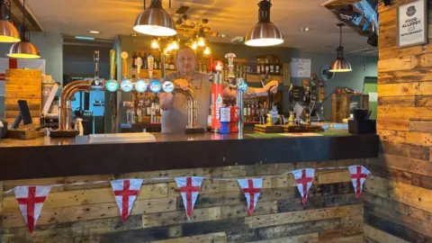 Jason Cleary A man stood behind a bar with England flags draped on a string.