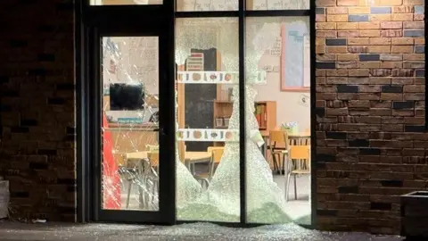 Glass doors and windows of a school which have been shattered. The rest of the wall is brick.