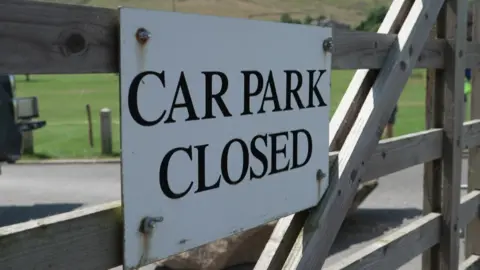 The image shows a white sign reading CAR PARK CLOSED attached to a wooden gate. Beyond the gate is a blurred rural landscape and a paved road.