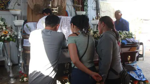 Two women and a man gaze at the open coffin of one of the victims of the shooting in Playas, Ecuador. Flowers have been placed on the closed part of the lid and the white lining of the coffin can be made out where it is open. In the background, there is a second coffin. 