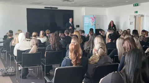 A man gives in a suit a speech at the front of a classroom. The backs of students heads are visable, sat in chairs in rows. A woman stands off to the side