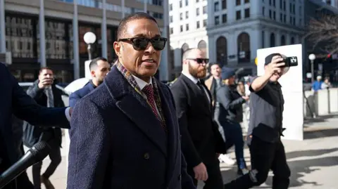 Don Lemon in sunglasses and a navy coat arrives in court, with a handful of media and security around him 