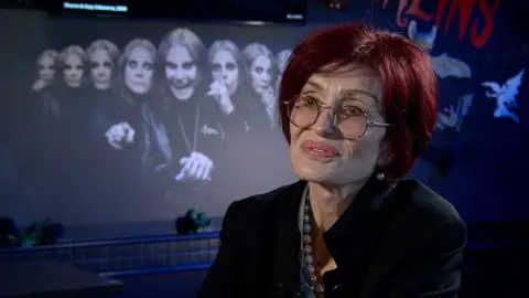 A woman is smiling off camera. Sharon Osbourne has red hair, thin rimmed glasses and red lips. Also wearing a black jacket and pearl necklace.
