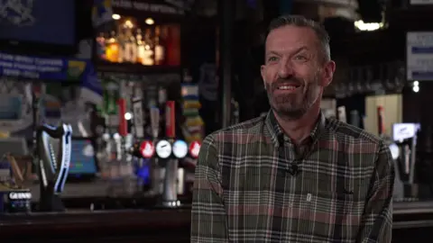 David Bond, who has short brown hair and brown stubble and wears a green checked shirt, smiles at the camera in front of the bar.