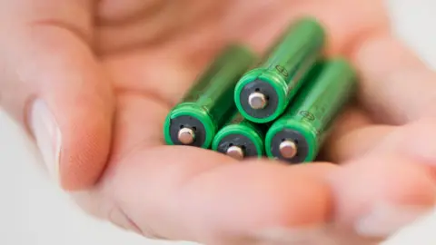 A hand holding four green coloured AA batteries.