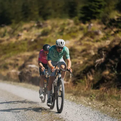Red On Sports Two riders pedal across the gravel with trees in the background