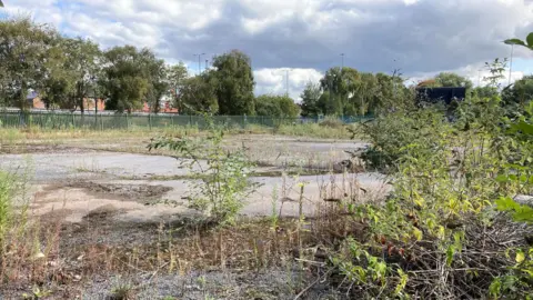 The picture shows the land at Phoenix Street in Derby which used to be a car park. It's overgrown with weeds and has broken tarmac. The land has a fence around it. 