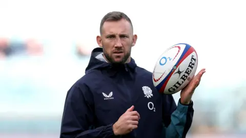 In an England-embroidered zip-up rain jacket, Byron McGuigan balances a rugby ball in his left hand as he watches on before a match