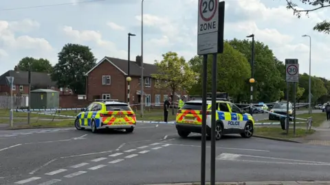 Two police cars sit in the middle of the road, which is taped off. Police officers can be seen in the background.