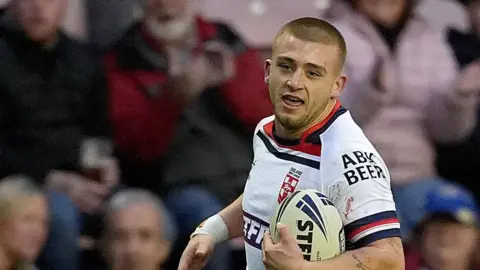 PA Media Mikey Lewis has cropped hair and is wearing a white Hull KR rugby shirt. He is running on the pitch with a rugby ball in front of spectators.