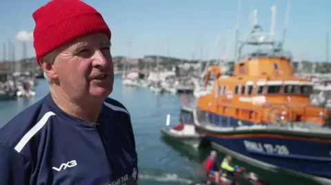 Richard Fowler is wearing a red wool hat and blue top and is standing by the orange and blue lifeboat in front of a harbour.