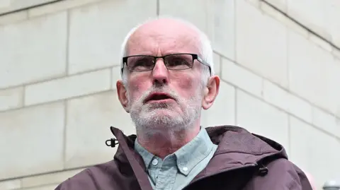 Noel McNally wearing black framed glasses a grey shirt and a black coat.