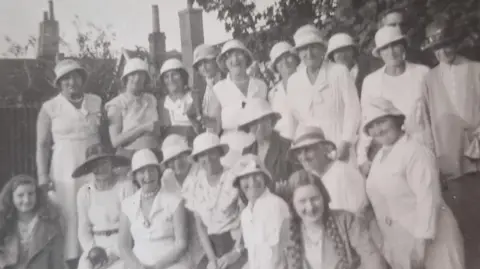A black and white image of a group of women wearing white bowler hats and white dresses. They are smiling at the camera. 