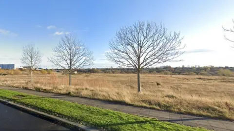 The large land off Cygnet Drive in Stockton is surrounded by several buildings in the distance. A number of young trees a re growing between the grassland and the pavement.