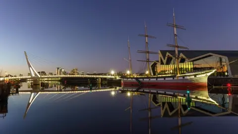 Ian Camrass Landscape of the River Clyde at the Transport museum with a boat moored outside and a bridge illuminated over the water below a darkening sky 