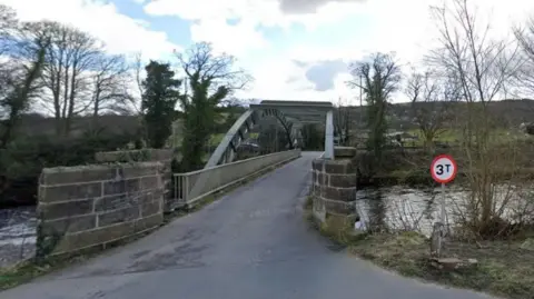 Google A bridge over a river. It is a metal bridge with railings on either side and moors and trees in the background. There is a sign which says 3T.