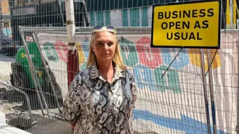Julie Goodwin has long blonde hair and has sunglasses on top of her head. She is wearing a grey and white patterned shirt and is standing in from of metal roadworks fencing. There is a yellow sign with black writing which reads Business open as usual.