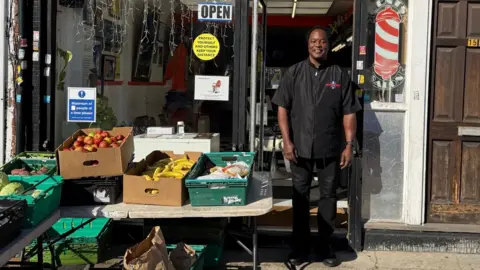 Ziggy Myers stands outside his barber shop. Next to him is a stand displaying boxes of fruit, including red apples, yellow banana and other items. Ziggy is wearing a black top and black trousers.