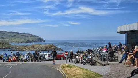 The Sound car park on a sunny day. It is busy with many bikes, cars and people. The Calf of Man is in the background with the sea in between, and the cafe is on the right.