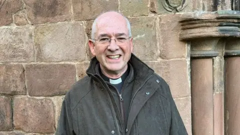 A male reverend, smiling and wearing clear-framed glasses, standing in front of a stone wall. 