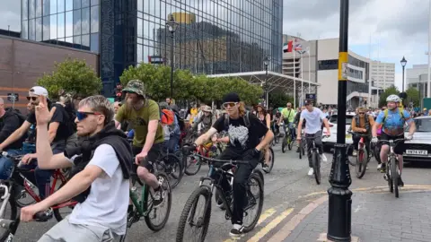 A group of men are cycling on their bikes, some are wearing helmets and glasses. They are dancing along to music as they cycle on a road. 