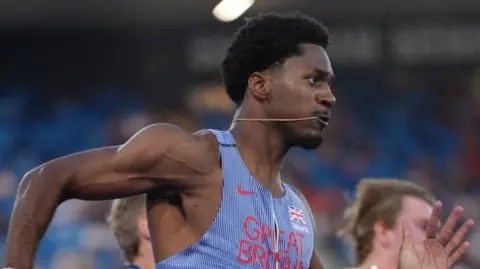 Ebuka Nwokeji wearing a blue and white Great Britain team vest while sprinting. He has black hair and a thin gold chain in his mouth.