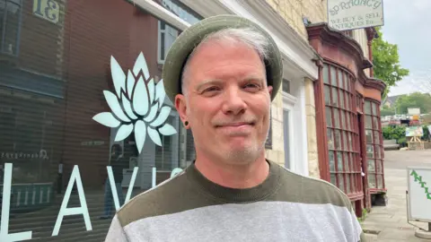 A photo showing a white man with grey hair and wearing a green hat and a green and grey jumper. He is stood on a street in front of a shop widnow