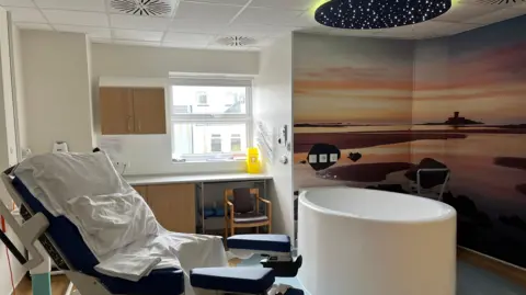 Inside a birthing room at the General Hospital in St Helier, Jersey. There is a hospital chair, a round bath, a sunset image on the wall and a window at the back.