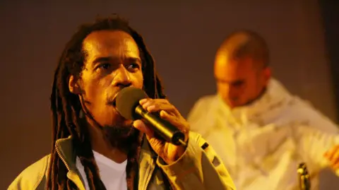 A man speaks into a microphone. He is wearing a white top and a green jacket, and he has long dreadlocks. He is standing under yellow lighting and another performer can be seen in the background.
