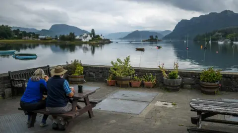 A woman and a man sit at a picnic bench with a view from Plockton of a loch and distant hills and mountains.
