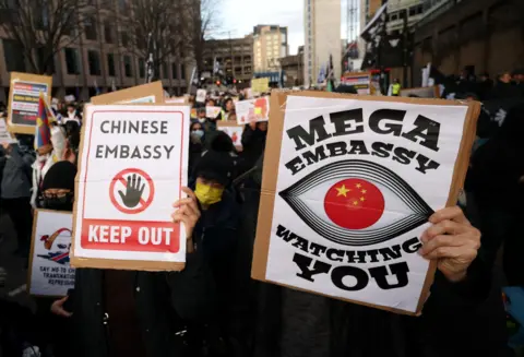 A large crowd of people gather along a road in the City of London, many holding up signs. Two people n the foreground hold up large signs reading 'Chinese Embassy - keep out' and 'Mega Embassy watching you'.
