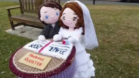 Carol Clayton A knitted pair of characters, who are wearing wedding garments, sign a register. The knitted item is placed on top of a parking bollard as a decoration.