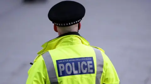 A Greater Manchester Police officer wearing a yellow high vis jacket and a hat faces away from the camera