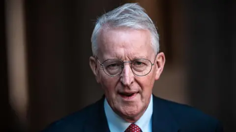 Getty A white haired man with round glasses, a navy suit, blue shirt and red and white polko-dot tie.