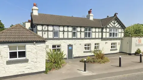 Google A two-storey white painted brick building with grey wood features and a pitched roof on one end.