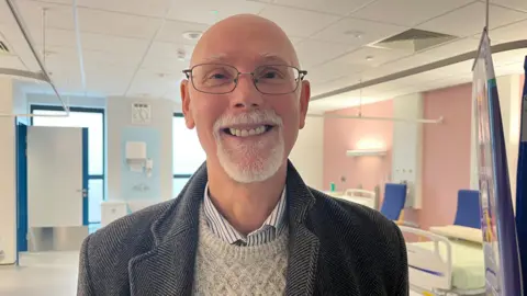 ELLEN KNIGHT/BBC Nick pictured smiling and looking into the camera. He's wearing a blue and white striped collared shirt, a cream cable-knit jumper and a dark grey wool herringbone coat. He's stood in a bay with two hospital beds visible over his left shoulder, which have green sheets and blue armchairs next to them. The walls of the room are light pink, and the floor and ceiling are white. 